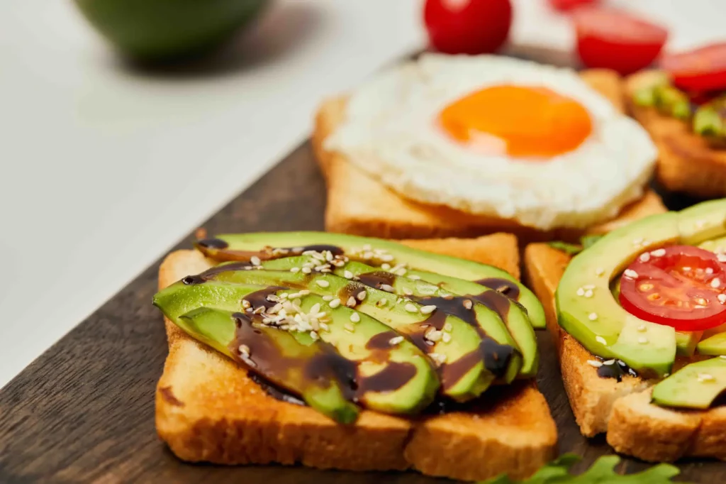 Nahaufnahme eines Frühstückstellers mit Toastscheiben, belegt mit Avocadoscheiben, einem Schuss Balsamico-Glasur, Sesamkörnern, Tomatenscheiben.