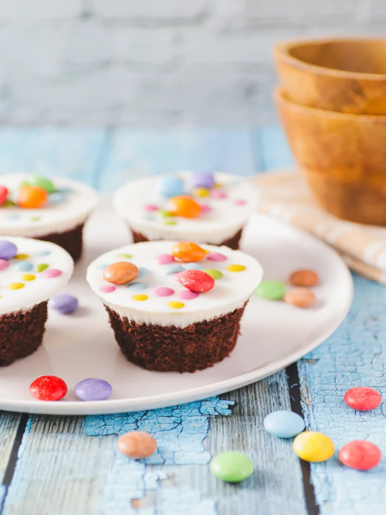 Ein weißer Teller mit vier Schokoladen-Cupcakes, garniert mit weißem Zuckerguss und bunten Bonbons.