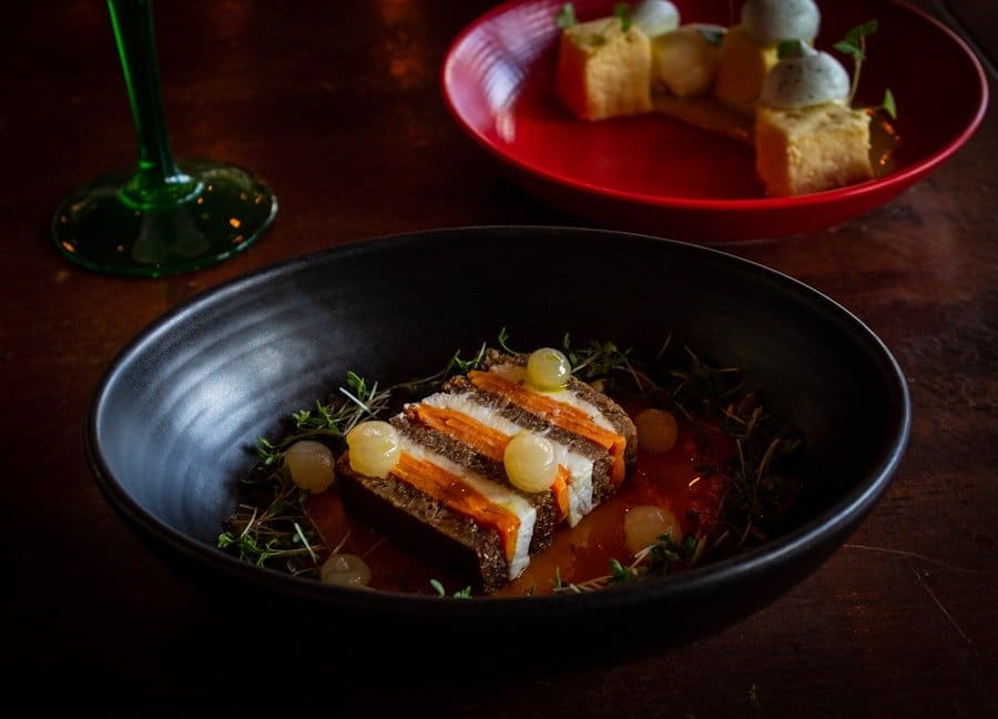 Der Food Fotograf - In einer schwarzen Schüssel liegt ein Teller mit Fischschichten und Orangensauce, garniert mit kleinen runden Gemüse- oder.