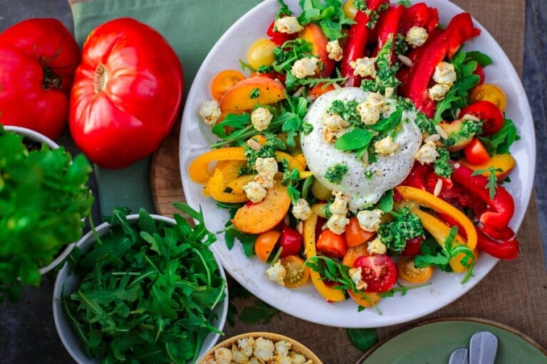 Food Fotograf Ein bunter Salat auf einem weißen Teller besteht aus Burrata-Käse, Rucola, roten und gelben Paprikaschoten, Kirschtomaten und Popcorn.