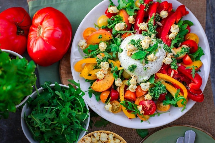 Food Fotograf Ein bunter Salat auf einem weißen Teller besteht aus Burrata-Käse, Rucola, roten und gelben Paprikaschoten, Kirschtomaten und Popcorn.