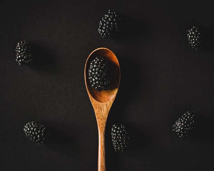 Der Food Fotograf - Auf einem Holzlöffel liegt eine einzelne Brombeere, umgeben von fünf weiteren Brombeeren, die auf einem schwarzen Hintergrund.