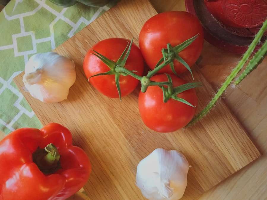 Drei rote Tomaten an einer Ranke, zwei Knoblauchknollen, eine rote Paprika und eine grüne Artischocke sind auf einem hölzernen Schneidebrett.