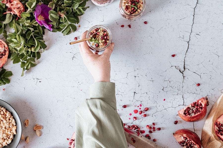 Der Food Fotograf - Eine Hand in einem grünen Langarmshirt hält ein Glas mit Müsli und Granatapfelkernen auf einer rissigen weißen Oberfläche.