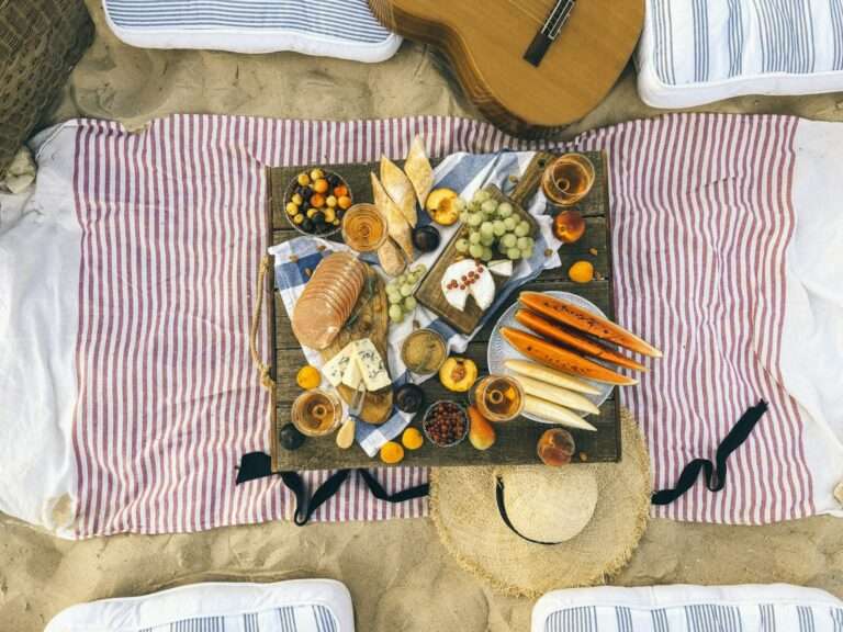 Ein Picknick-Setup im Sand umfasst ein Holztablett mit verschiedenen Käsesorten, Brot, Trauben, Pfirsichen, Wassermelonenscheiben und Nüssen.