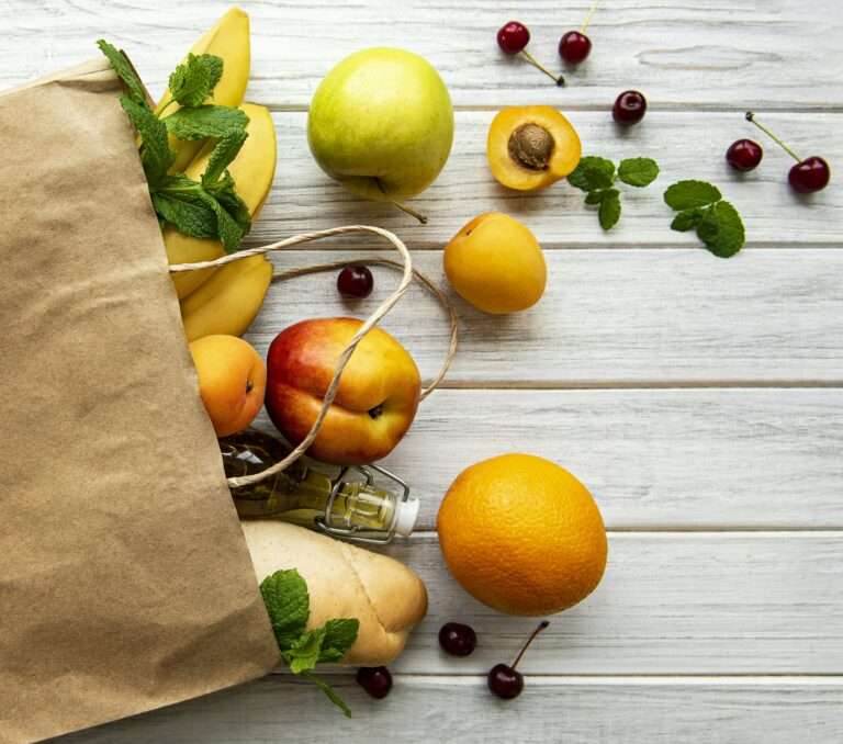 Eine Papiertüte liegt teilweise geöffnet auf einer weißen Holzoberfläche und gibt den Blick frei auf Bananen, Aprikosen, Äpfel und einen Pfirsich.