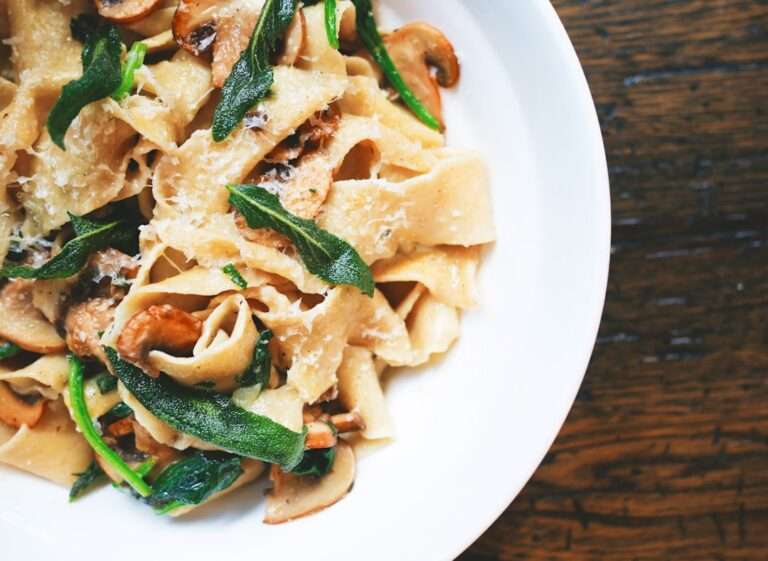 Der Food Fotograf - Ein weißer Teller mit Pappardelle-Nudeln ist mit sautierten Pilzen, frischem grünem Gemüse und geriebenem Käse garniert.