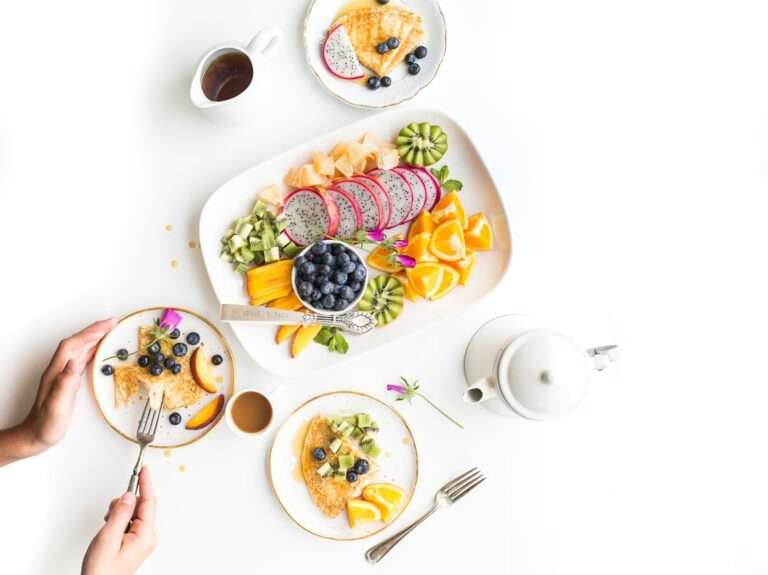 Der Food Fotograf - Eine Person isst eine Mahlzeit mit zwei Tellern Pfannkuchen, belegt mit Kiwi, Blaubeeren und Orangenscheiben.