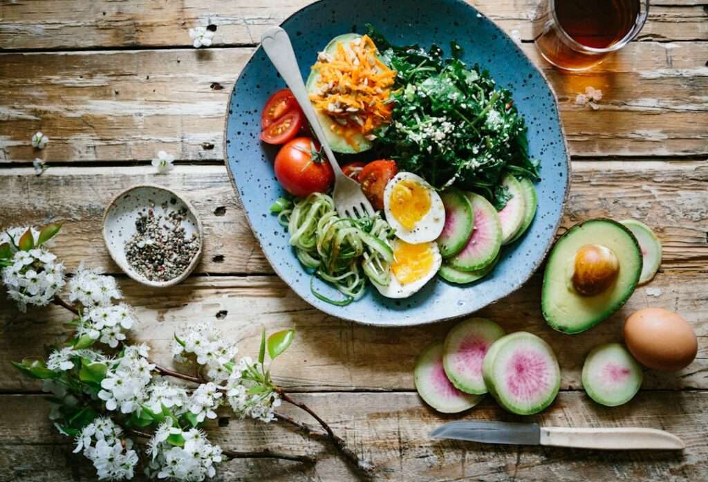 Auf einem rustikalen Holztisch steht ein blauer Teller mit einer Mischung aus Salatgemüse, darunter Blattgemüse, Zucchini-Nudeln, Avocadoscheiben.
