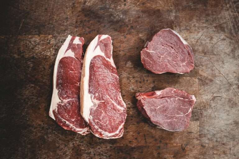 Ein Holztisch mit zwei Stücken rohem Ribeye-Steak links und zwei Stücken rohem Filet-Mignon-Steak rechts, perfekt als Hintergrund für Food-Fotografie.