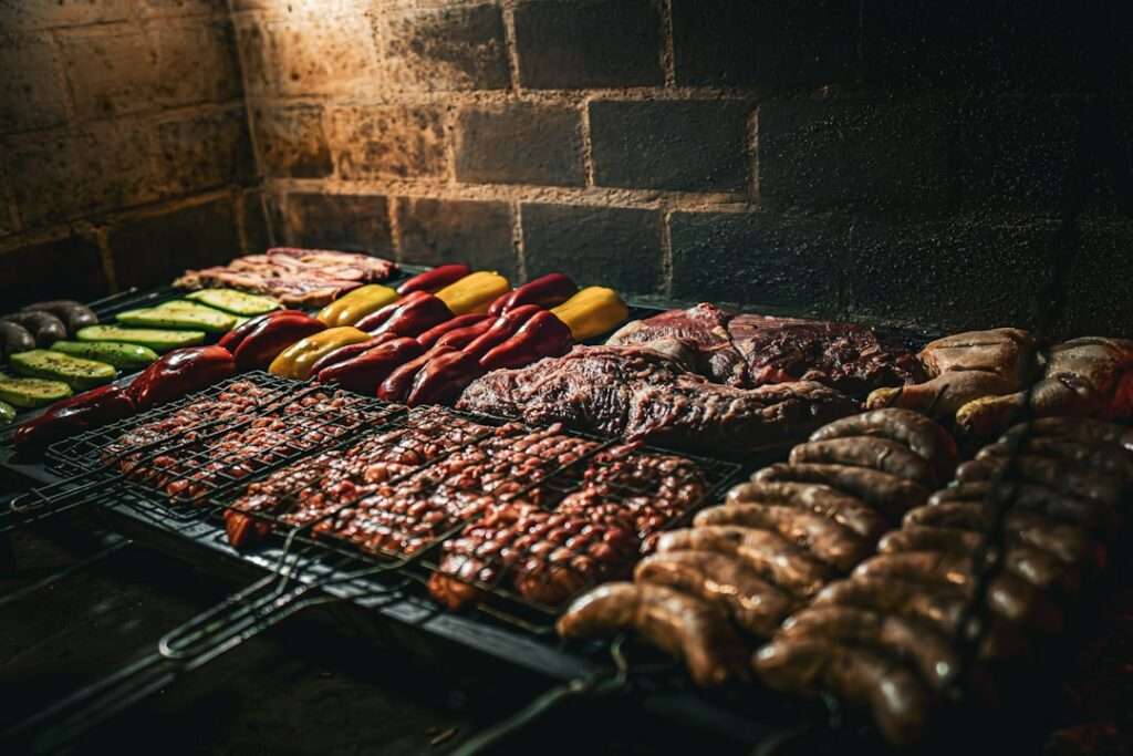 Verschiedene Fleischsorten, Wurst und Gemüse, darunter Paprika und Zucchini, werden über offener Flamme gegrillt.