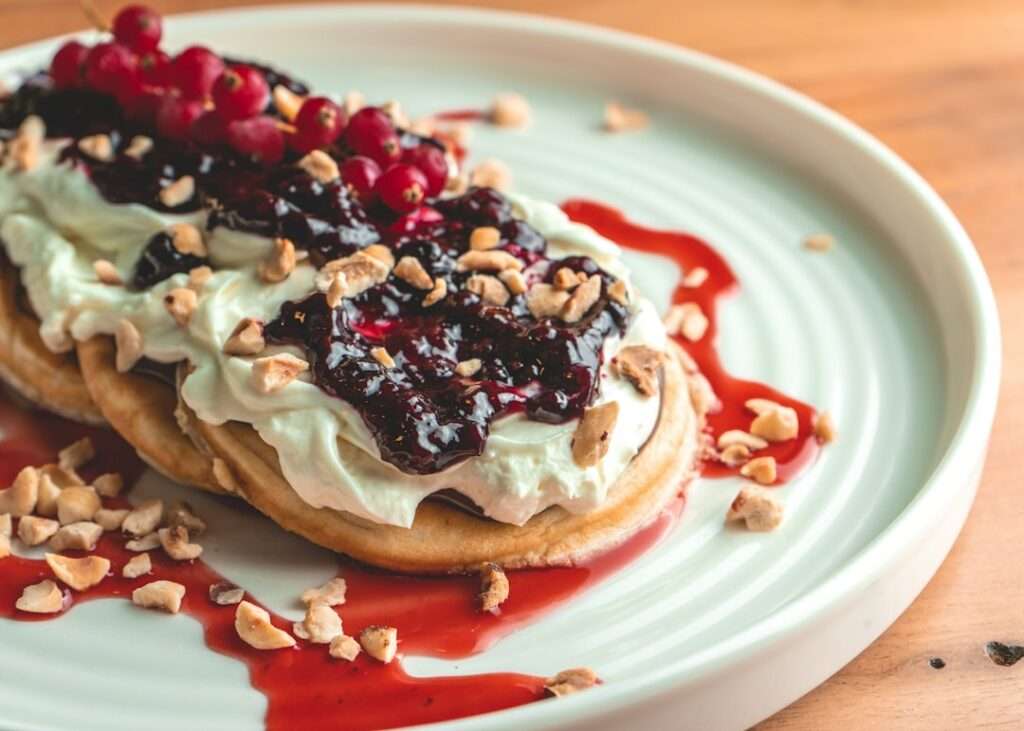 Der Food Fotograf - Auf einem Teller liegt ein Dessert mit Gebäck, das mit Schlagsahne und dunkler Beerensauce belegt und mit gehackten Nüssen.