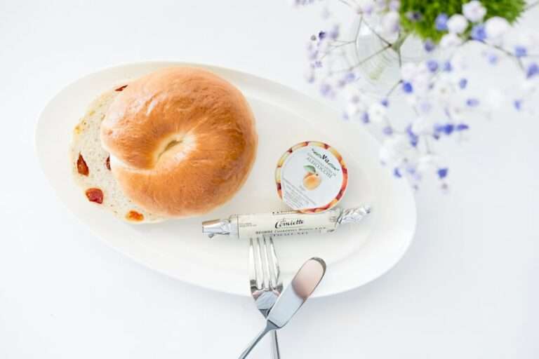 Der Food Fotograf - Ein Teller mit einem halbierten Bagel, einem kleinen Behälter mit Aprikosenmarmelade und einem eingewickelten Stück Käse.