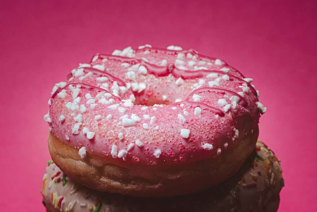Eine Nahaufnahme eines Donuts mit rosa Zuckerguss, weißen Streuseln und einem Spritzer rosa Zuckerguss obendrauf, vor einem rosa Hintergrund.