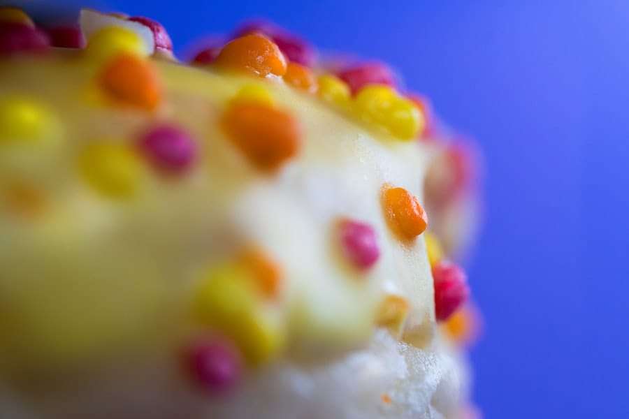 Nahaufnahme eines Desserts mit cremeweißem Belag und bunten runden Streuseln, darunter Orange, Gelb und Rosa, vor einem verschwommenen blauen.