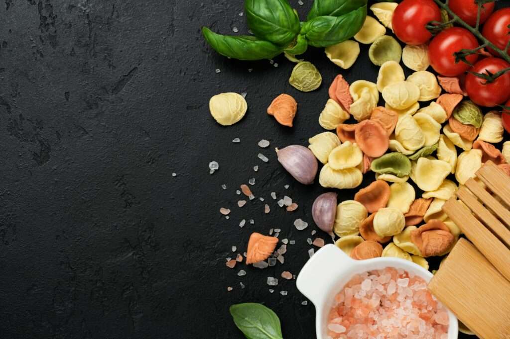 Eine Auswahl trockener, bunter Tortellini, Kirschtomaten, Knoblauchzehen, frische Basilikumblätter und ein Holzlöffel sind auf einer dunklen.