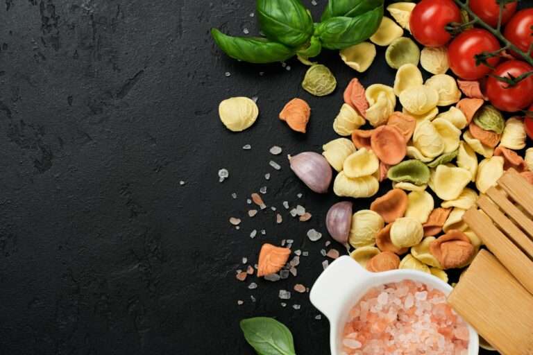 Eine Auswahl trockener, bunter Tortellini, Kirschtomaten, Knoblauchzehen, frische Basilikumblätter und ein Holzlöffel sind auf einer dunklen.