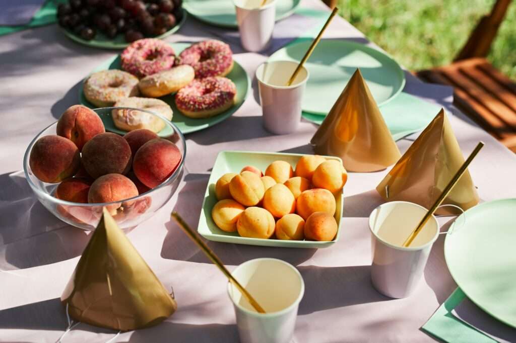 Ein für eine Party gedeckter Tisch. Es gibt Schalen mit Pfirsichen, Teller mit Donuts, Pappbecher mit Strohhalmen und goldene Partyhüte.