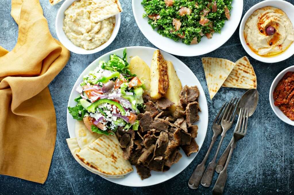 Ein Teller mit mediterranem Essen umfasst Gyrosfleisch vom Lamm, einen griechischen Salat mit Feta, Pita-Brot, Kartoffelecken und Dip-Saucen.