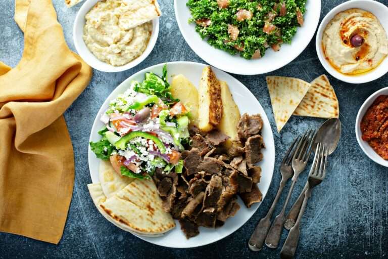 Ein Teller mit mediterranem Essen umfasst Gyrosfleisch vom Lamm, einen griechischen Salat mit Feta, Pita-Brot, Kartoffelecken und Dip-Saucen.