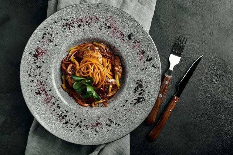 Ein Nudelgericht mit Spaghetti, Fleisch und Garnitur auf einem strukturierten, grauen Teller steht auf einem grauen Tuch.