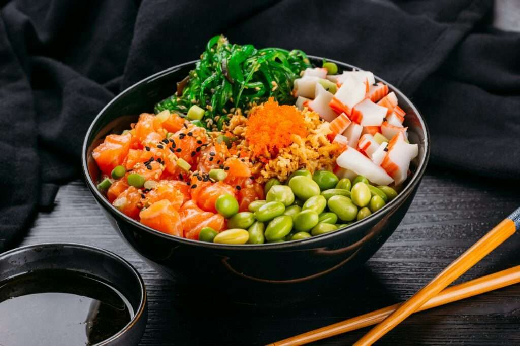 Eine Schüssel mit Sushi-Zutaten enthält gewürfelten Lachs, Edamame, Seetangsalat, Krabbenstäbchen, Sesamsamen und orangefarbenen Fischrogen.