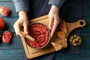 Eine Person taucht ein Stück Brot in eine Schüssel mit Rote-Bete-Hummus auf einem rustikalen Holzbrett und perfektioniert damit ihr.