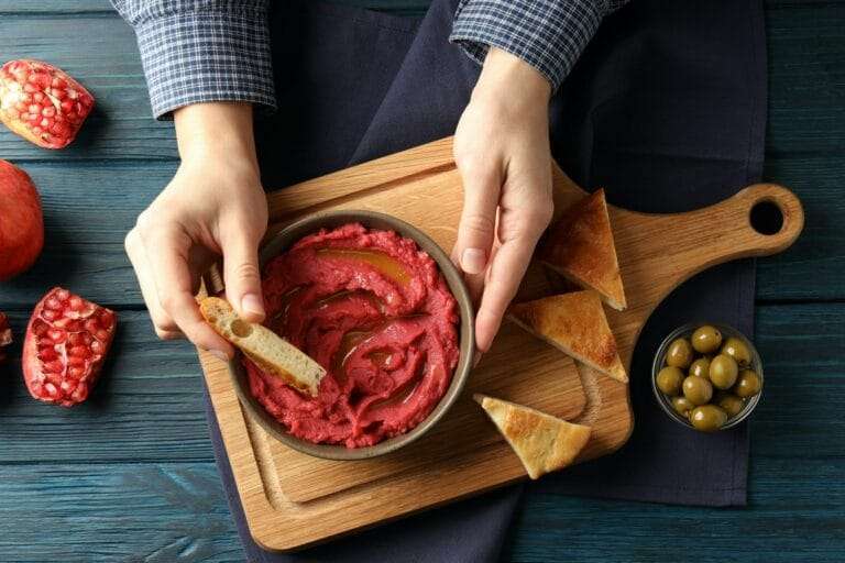 Eine Person taucht ein Stück Brot in eine Schüssel mit Rote-Bete-Hummus auf einem rustikalen Holzbrett und perfektioniert damit ihr.