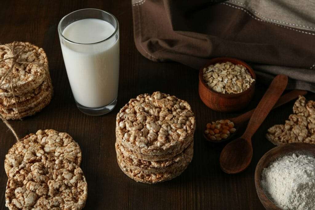 Ein Holztisch mit Reiskuchen, einem Glas Milch, kleinen Schüsseln mit Haferflocken und Maiskörnern und einem Holzlöffel mit Mehl.