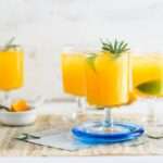 Vier Gläser Orangensaft mit Limettenscheiben und Rosmarinzweigen stehen auf einem Tisch.