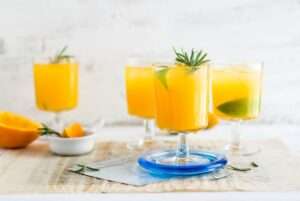 Vier Gläser Orangensaft mit Limettenscheiben und Rosmarinzweigen stehen auf einem Tisch.