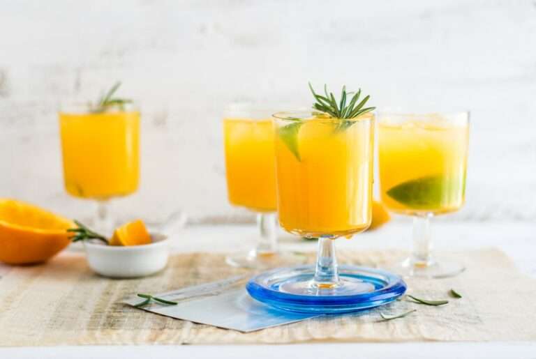 Vier Gläser Orangensaft mit Limettenscheiben und Rosmarinzweigen stehen auf einem Tisch.