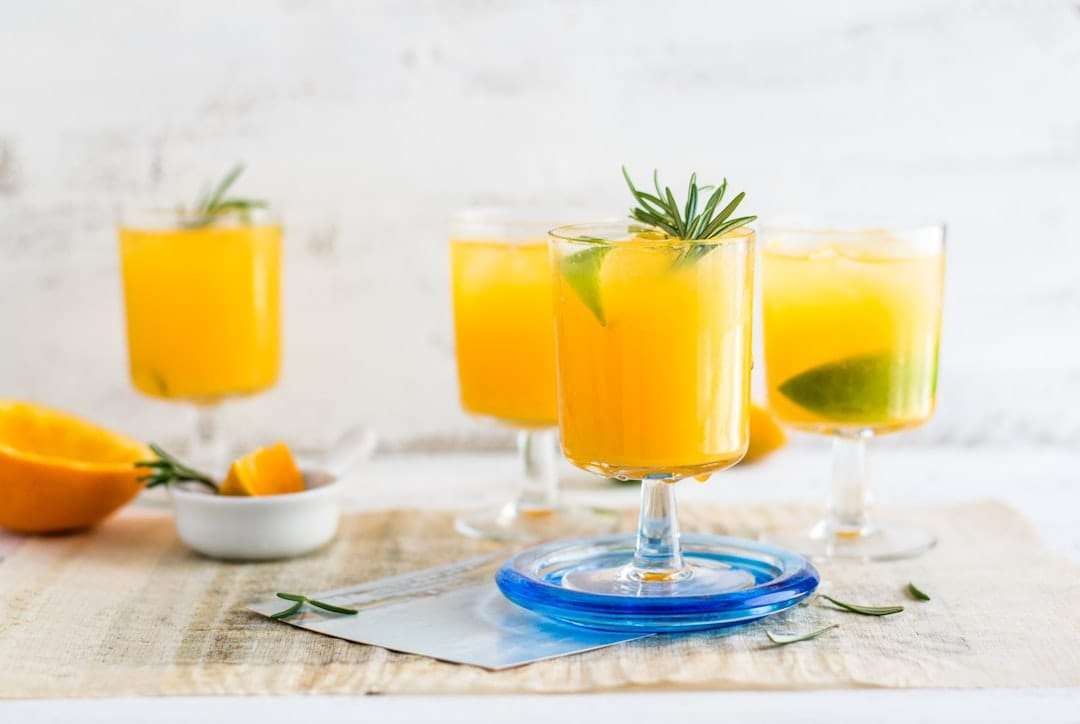Vier Gläser Orangensaft mit Limettenscheiben und Rosmarinzweigen stehen auf einem Tisch.