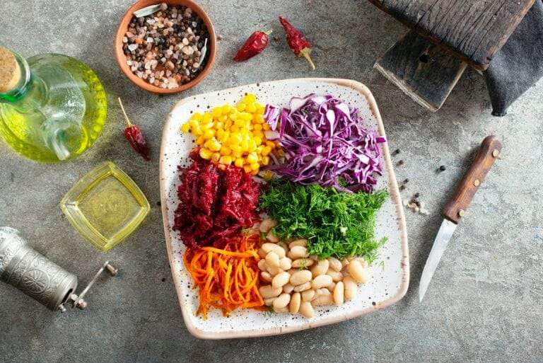 Ein Teller mit verschiedenen Gemüsesorten, darunter Mais, zerkleinerter Rotkohl, geriebene Rote Bete, geriebene Karotten, Dill und weiße Bohnen.