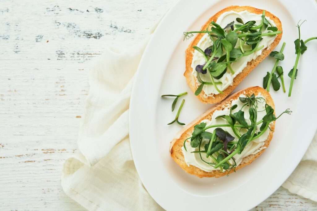 Zwei Brotscheiben mit Frischkäse, Gurkenscheiben und einer Mischung aus Microgreens sind kunstvoll auf einem weißen ovalen Teller arrangiert.