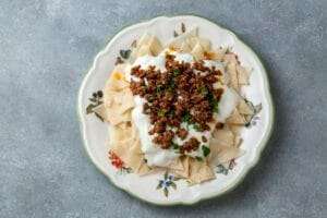 Ein Teller mit traditionellem türkischem Mantı, belegt mit Joghurt und gewürztem Hackfleisch, steht auf einem Teller mit Blumenmuster.