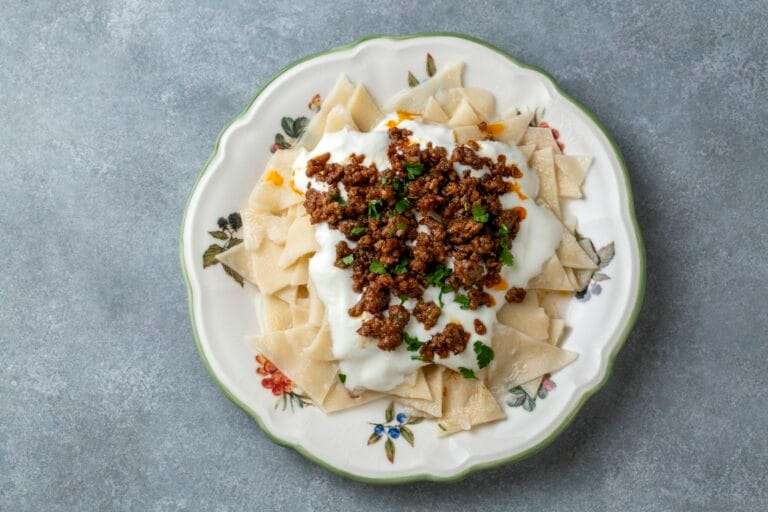 Ein Teller mit traditionellem türkischem Mantı, belegt mit Joghurt und gewürztem Hackfleisch, steht auf einem Teller mit Blumenmuster.
