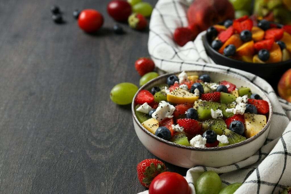 Eine Schüssel Obstsalat mit Erdbeeren, Blaubeeren, Kiwi, Trauben und Käsestreuseln steht auf einer dunklen Holzoberfläche – perfekt für jede Art.