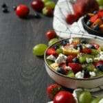 Eine Schüssel Obstsalat mit Erdbeeren, Blaubeeren, Kiwi, Trauben und Käsestreuseln steht auf einer dunklen Holzoberfläche – perfekt für jede Art.