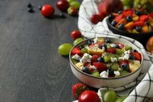 Eine Schüssel Obstsalat mit Erdbeeren, Blaubeeren, Kiwi, Trauben und Käsestreuseln steht auf einer dunklen Holzoberfläche – perfekt für jede Art.
