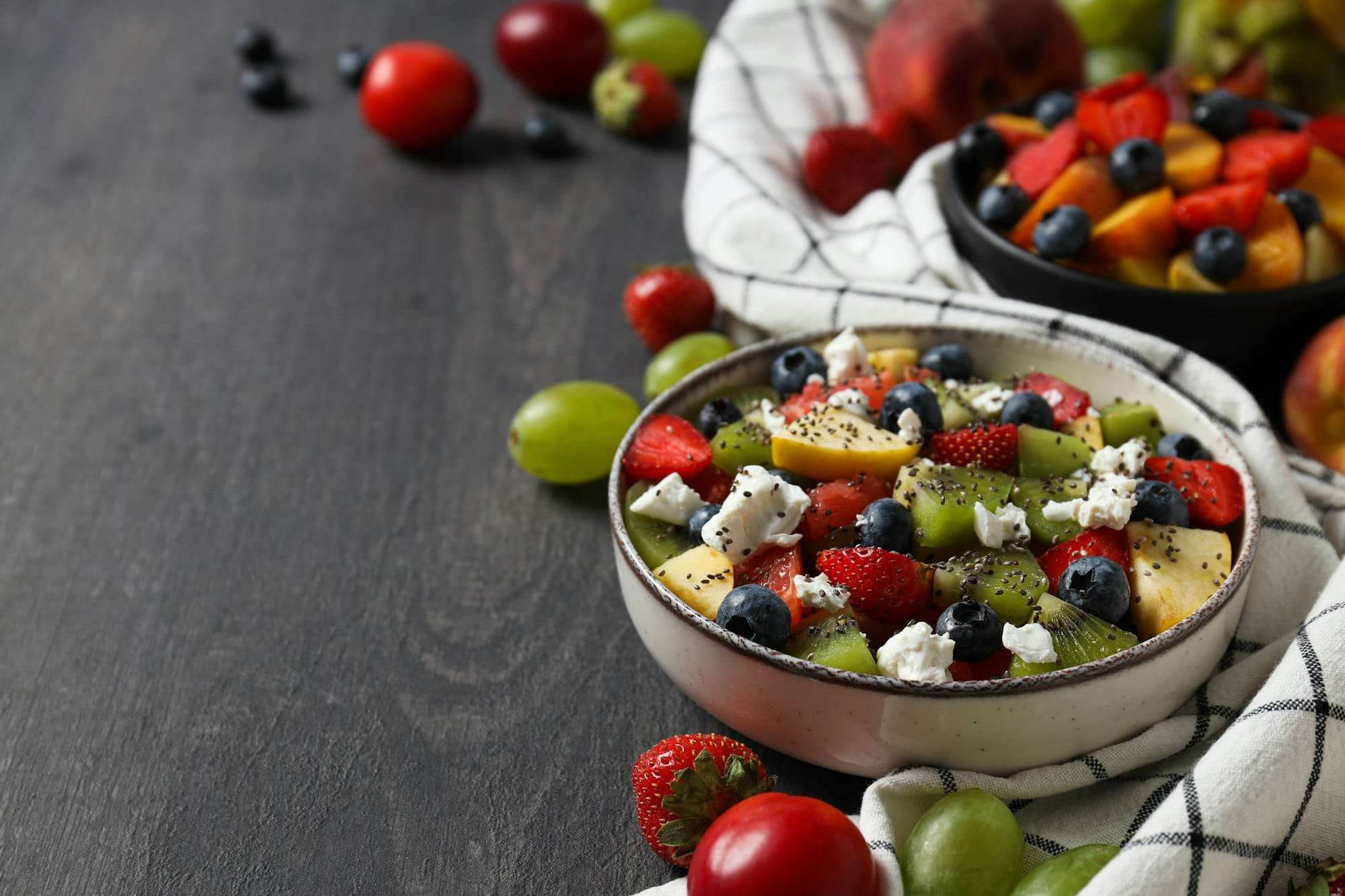 Eine Schüssel Obstsalat mit Erdbeeren, Blaubeeren, Kiwi, Trauben und Käsestreuseln steht auf einer dunklen Holzoberfläche – perfekt für jede Art.